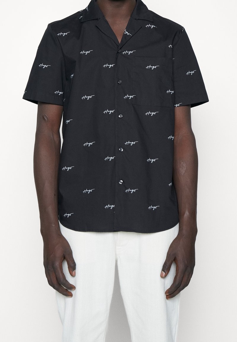 Zwart T-shirt met korte mouwen, voorzien van witte schriftprint, knoopsluiting aan de voorkant, liggende kraag en een enkele borstzak, gemaakt van een lichtgewicht stof.