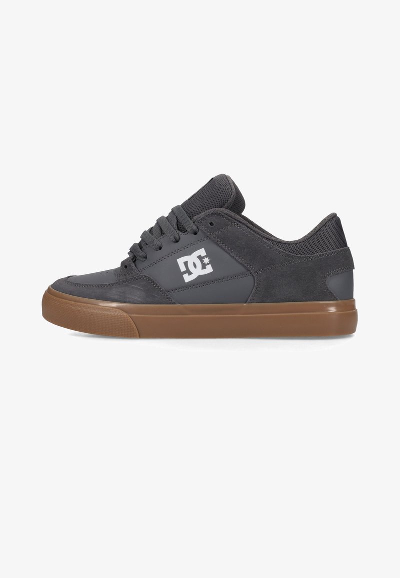 Chaussure de skate basse gris foncé avec semelle en caoutchouc marron, lacets noirs et logo DC blanc sur la tige latérale.
