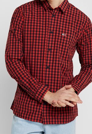 Man draagt een rood-zwart geruit overhemd met knopen en lange mouwen, gecombineerd met lichtblauwe spijkerbroek, handen in elkaar gevouwen ter hoogte van de taille.