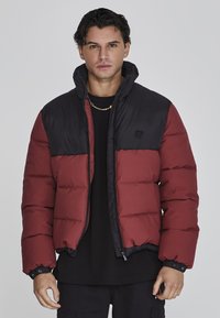 Chaqueta acolchada para hombre con una sección superior negra y un fondo burdeos. Material ligero, textura acolchada y puños ajustables. Detalle del logo en negro.
