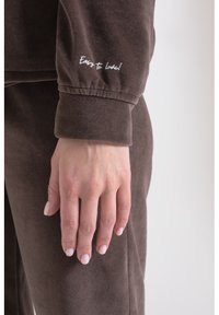 Brązowy, miękki materiałowy strój do relaksu z prążkowanymi mankietami. Na rękawie znajduje się biały napis "Łatwy do kochania!", podkreślający wygodę i swobodny styl.