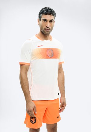 Uomo che indossa divisa da calcio dei Paesi Bassi bianca e arancione con stemma del leone sulla maglia e sui pantaloncini, in piedi contro uno sfondo chiaro e uniforme.