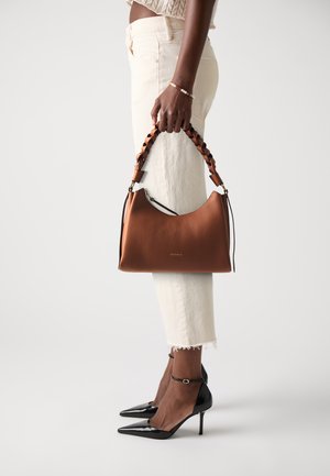 Model wearing cropped light beige jeans and black high heels holding a brown leather handbag with twisted handle by their side.