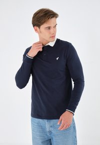 Marineblaues Langarm-Polohemd mit weißem Kragen und Manschetten, das ein kleines weißes Vogel-Logo auf der Brust und eine Knopfleiste aufweist.