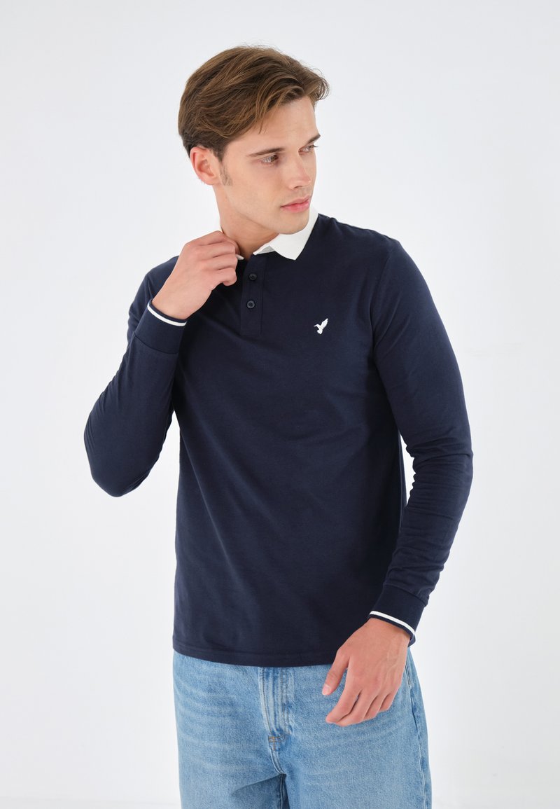 Marineblaues Langarm-Polohemd mit weißem Kragen und Manschetten, das ein kleines weißes Vogel-Logo auf der Brust und eine Knopfleiste aufweist.