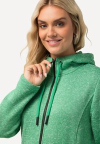 Grön hoodie i texturerad tyg med dragkedja fram, svarta detaljer och kontrasterande dragsnören. Nära bild som visar hand på dragkedjan.