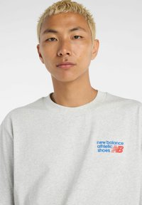 Grå sweatshirt med rund halsringning. Har en blå och röd logotyp på vänster bröst: "new balance athletic shoes" tillsammans med ett märke. Mjuk textur.