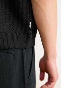 Černý pletený svetr s texturovaným vzorem, s žebrovaným lemem a malou bílou štítkem „OBEY“ na boku, doplněný tmavými kalhotami.