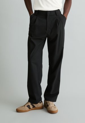 Homme portant un pantalon plissé noir et des baskets beiges avec des bandes noires, debout les mains dans les poches sur un fond neutre.