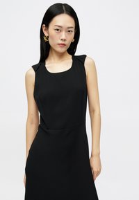 Νέα γυναίκα με ίσια μαύρα μαλλιά, φορώντας χρυσά αφηρημένα σκουλαρίκια και ένα αμάνικο μαύρο φόρεμα, σε φόντο λευκό.