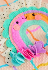 Decoración de fieltro colorida con un diseño sonriente de arcoíris y flores superpuestas en tonos rosas y púrpuras, sobre un fondo pastel texturizado.