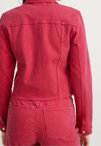 Červená džínová bunda s přiléhavým střihem, viditelným šitím a cvoky. Má zkrácenou délku a roztrhaný lem.