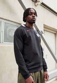 Sudadera negra con cuello redondo, que presenta un logo azul y acentos rojos. Textura suave, combinada con pantalones de color verde oliva y puños acanalados.