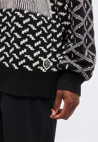 Gros plan d'une personne portant un pull noir et blanc à motifs géométriques avec des poignets côtelés, montrant sa main sur le côté.