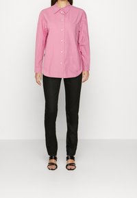 Camicia rosa a maniche lunghe con colletto, dotata di taschino sul petto, abbinata a pantaloni neri a gamba dritta e sandali neri con cinturini.