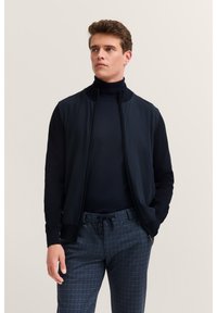 Granatowa kurtka bombowiec z zamkiem z przodu, dzianinowym kołnierzem i ciemnymi rękawami, zestawiona z strukturami w kratę w odcieniach niebieskiego.