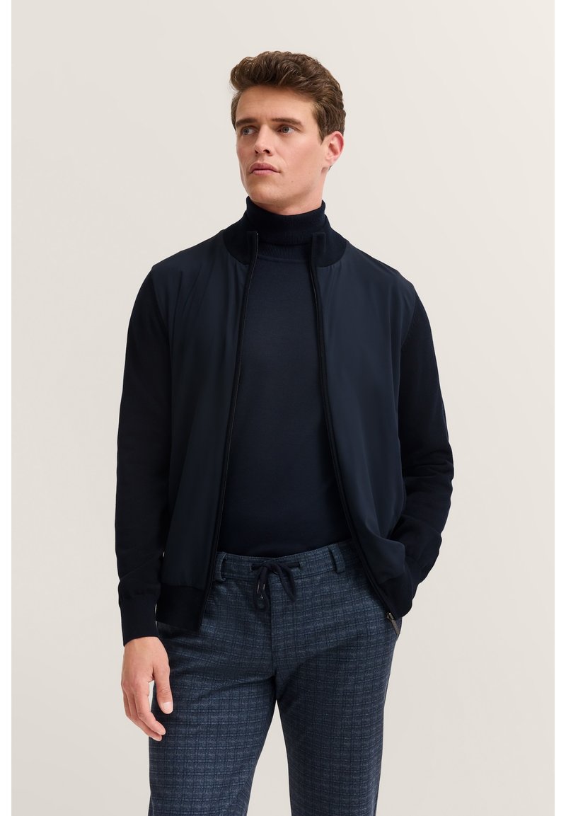 Granatowa kurtka bombowiec z zamkiem z przodu, dzianinowym kołnierzem i ciemnymi rękawami, zestawiona z strukturami w kratę w odcieniach niebieskiego.