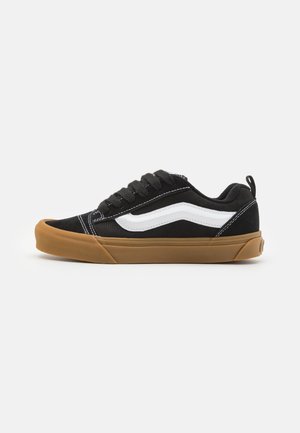 Sort lavtop sneaker med hvid side-stribe, brun gummisål, sorte snørebånd og trækstrop på hælen, vist i profil mod en hvid baggrund.