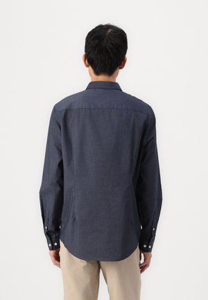 Homme de dos portant une chemise bleu foncé à manches longues boutonnée et un pantalon beige sur un fond blanc uni.