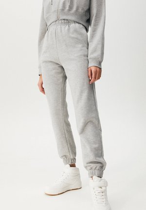Person iført lysegrå joggers med elastiske manchetter og hvide høje sneakers, stående mod en ensfarvet baggrund.