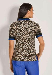 Vrouw die wegdraait, gekleed in een korte mouwen shirt met luipaardprint met zwarte en blauwe accenten en felblauwe broek, staande tegen een effen muur.