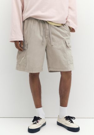 Muž v béžových cargo šortkách so sťahovacou šnúrkou, bielych ponožkách a krémových teniskách s čiernymi podrážkami, stojaci na bielom pozadí.