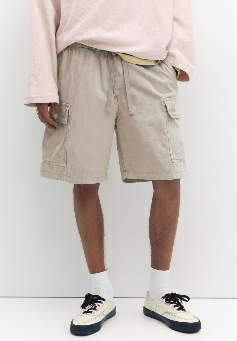 Homme portant un short cargo beige avec cordon, des chaussettes blanches et des baskets crème avec des semelles noires, debout sur un fond blanc.