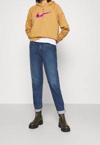 Hořčicově žlutá mikina s růžovým logem Nike, v kombinaci s modrými denimovými džíny ohrnutými na manžetách a tmavě hnědými kotníkovými botami s mohutnou podrážkou.