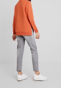 Sudadera de manga larga naranja con dobladillo curvado, combinada con jeans gris ajustados y zapatillas blancas, que muestran un estilo casual y materiales.