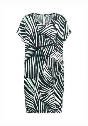 Femme marchant portant une robe mi-longue au motif abstrait noir et blanc et des chaussures plates beiges sur un fond beige uni.
