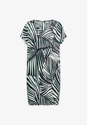 Femme marchant portant une robe mi-longue au motif abstrait noir et blanc et des chaussures plates beiges sur un fond beige uni.