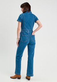 Jumpsuit in denim blu medio, maniche corte, con collo alla coreana, gamba a zampa d'elefante, due tasche posteriori e vita aderente.