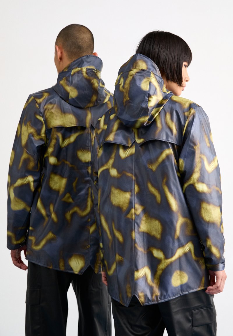 Veste étanche avec un motif abstrait jaune et noir, capuche et rabats à l'arrière, de coupe ample et texture lisse.