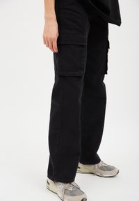 Schwarze Cargo-Hosen mit Seitentaschen, aus einem Baumwollmix gefertigt. Entworfen mit geradem Bein und verstärkten Nähten. Getragen mit hellfarbenen Sneakers.