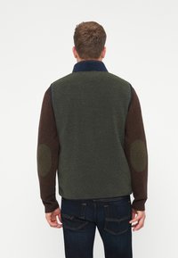 Vesta z fleece s farebným blokovaním: tmavozelené telo, hnedé rukávy a námornícky golier. Obsahuje lakťové záplaty a ribbované manžety.