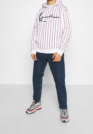 Bílá mikina s kapucí s tmavě červenými svislými pruhy, zdobená černým logem, v kombinaci s modrými džínami a černo-červenými teniskami.