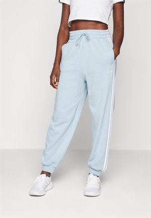 Persoon draagt lichtblauwe Adidas trainingsbroek met witte zijstrepen, gecombineerd met witte sneakers en een wit cropped top.