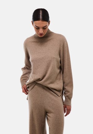 Un pull en tricot beige léger avec un col monté, une coupe décontractée et des manches longues, associé à un pantalon beige assorti. Texture douce.