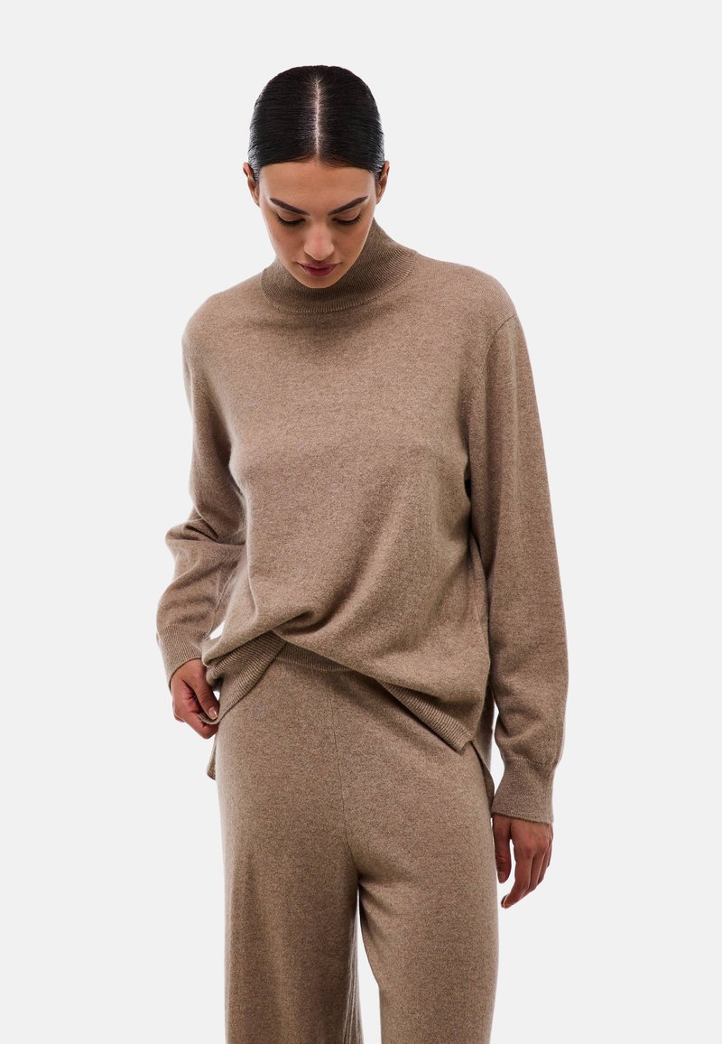 Un pull en tricot beige léger avec un col monté, une coupe décontractée et des manches longues, associé à un pantalon beige assorti. Texture douce.