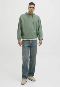 Grøn hættetrøje med lomme foran, sammen med lyseblå lige ben jeans og beige slip-on sko. Neutral baggrund.