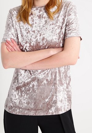 Personne portant un haut en velours écrasé à manches courtes de couleur mauve clair, bras croisés, associé à un pantalon noir sur un fond uni.