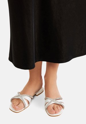 Bela sandala s ravnim peto, s prečnimi trakovi okrašenimi s srebrnimi dodatki. Utori na podplatu z minimalno teksturo.