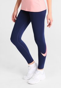 Kvinna som bär marinblå leggings med en rosa logotyp på vaden, en rosa topp, vita strumpor och vita träningsskor, stående mot en vit bakgrund.