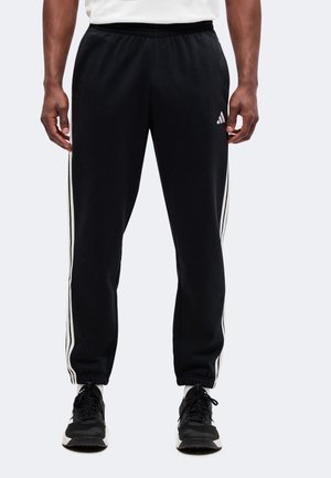 Άνδρας που φοράει μαύρο παντελόνι Adidas με άσπρες ρίγες στα πλάγια και μαύρα αθλητικά παπούτσια, στέκεται μπροστά σε απλό φόντο.