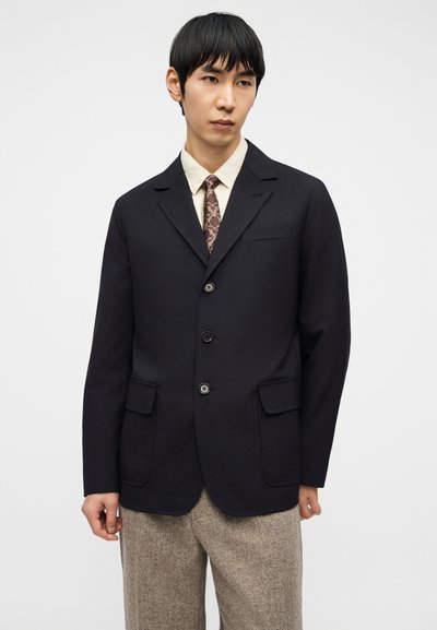 Άνδρας που φορά μαύρο σακάκι με τρία κουμπιά, γραβάτα με σχέδια, μπεζ πουκάμισο και υφασμάτινο μπεζ παντελόνι σε απλό φωτεινό φόντο.