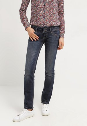 Osoba ubrana w wyblakłe, ciemnoniebieskie dżinsy, kwiecistą koszulę z długim rękawem i białe trampki, stojąca z jedną ręką na biodrze na białym tle.