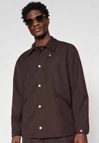 Brown, textured jacket with a collar, white snap buttons, and side pockets. Worn with dark tinted sunglasses. Simple design overall.