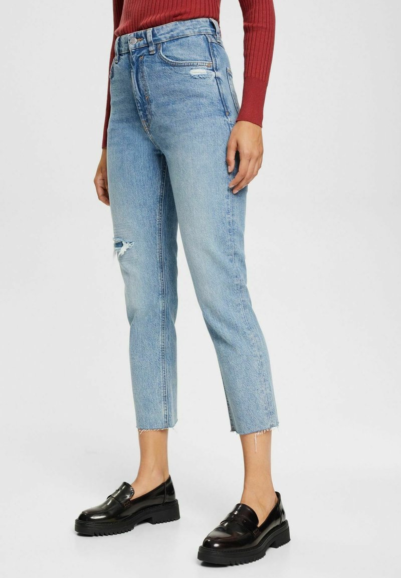 Personne portant un jean déchiré court bleu clair, des mocassins noirs épais et un haut à manches longues côtelé rouge, debout devant un fond uni.