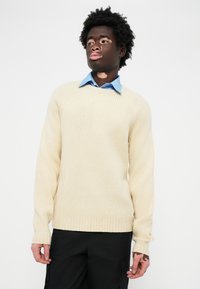 Cream ribbed sweater with long sleeves, round neck, and textured fabric, layered over a light blue collared shirt. Black pants visible.
