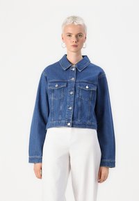 Jaqueta de denim cortada em azul médio, com colarinho aberto, bolsos frontais, botões de pressão prateados e mangas longas com um corte relaxado.